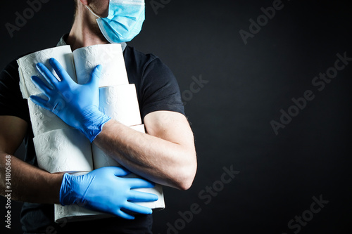 A young man dressed in a black T-shirt with blue rubber gloves and a protective mask on his face holds a pile of toilet paper in both hands. He's making a supply and prepares for a COVID-19 crisis.