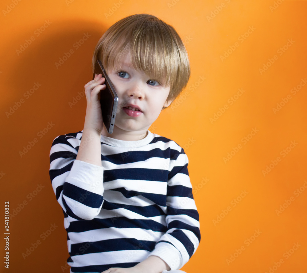 Little boy smiling, talking by smartphone, looking at side, posing against orange studio background. Happy childhood, gadget, advertising. Close up, copy space Little boy smiling, talking by smartphone, looking at side, posing against orange studio background. Happy childhood, gadget, advertising. Close up, copy space