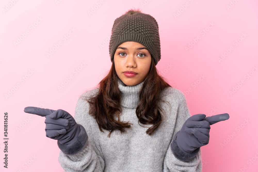 Young Colombian girl with winter hat over isolated pink wall pointing to the laterals having doubts Young Colombian girl with winter hat over isolated pink wall pointing to the laterals having doubts