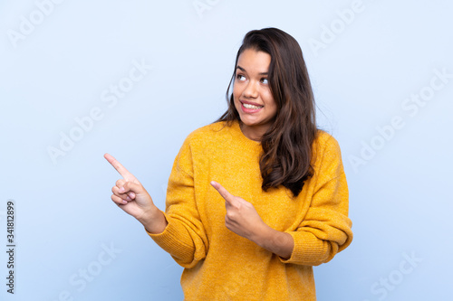 Young Colombian girl with sweater over isolated blue background frightened and pointing to the side