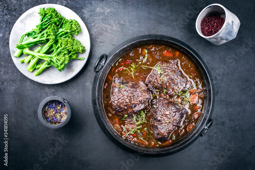 Traditional German braised beef cheeks in brown red wine sauce with carrots and broccoli offered as top view in a modern design stewpot on an old rustic board