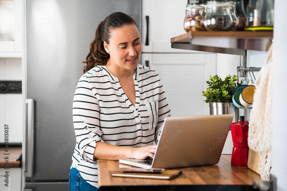 Smiling young woman working with laptop in her apartment kitchen. Working at home and stay at home. Smiling young woman working with laptop in her apartment kitchen. Working at home and stay at home.