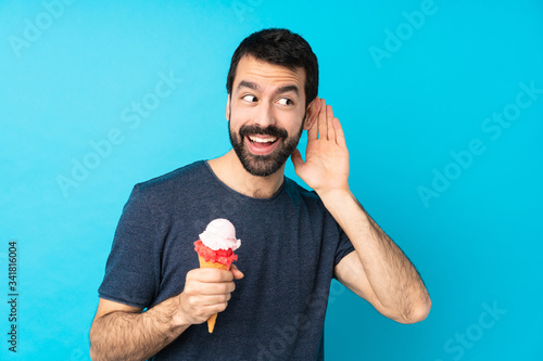 Young man with a cornet ice cream over isolated blue background listening to something by putting hand on the ear