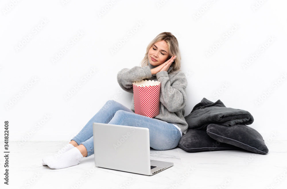 Teenager blonde girl eating popcorn while watching a movie on the laptop making sleep gesture in dorable expression Teenager blonde girl eating popcorn while watching a movie on the laptop making sleep gesture in dorable expression