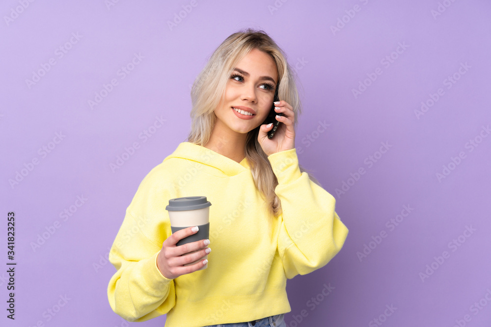 Teenager girl wearing a yellow sweatshirt over isolated purple background holding coffee to take away and a mobile Teenager girl wearing a yellow sweatshirt over isolated purple background holding coffee to take away and a mobile