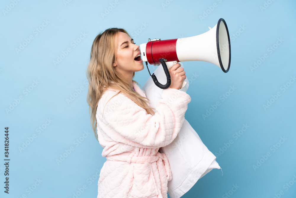 Teenager Russian girl in pajamas isolated on blue background shouting through a megaphone Teenager Russian girl in pajamas isolated on blue background shouting through a megaphone