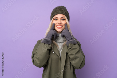 Teenager blonde girl with winter hat over isolated purple background smiling with a happy and pleasant expression