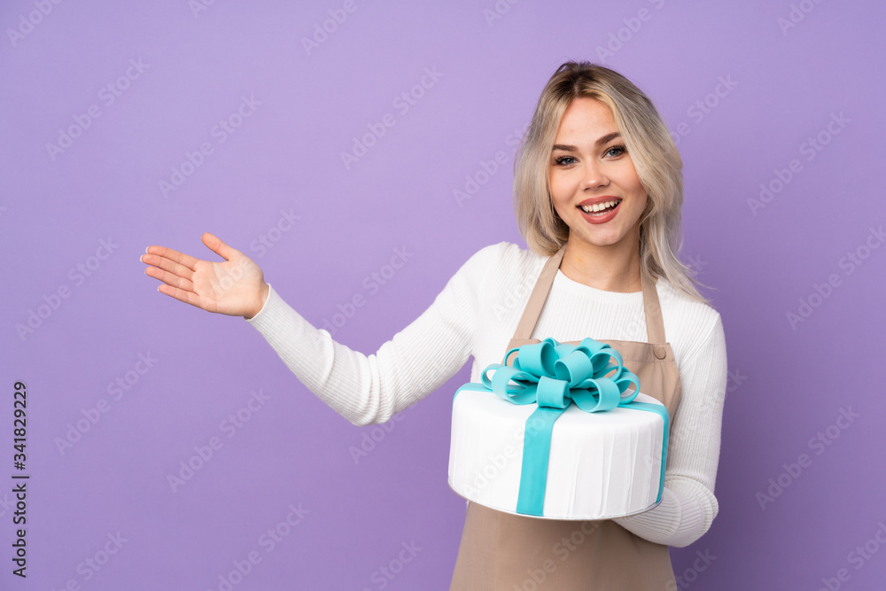 Young pastry chef holding a big cake over isolated purple background extending hands to the side for inviting to come Young pastry chef holding a big cake over isolated purple background extending hands to the side for inviting to come