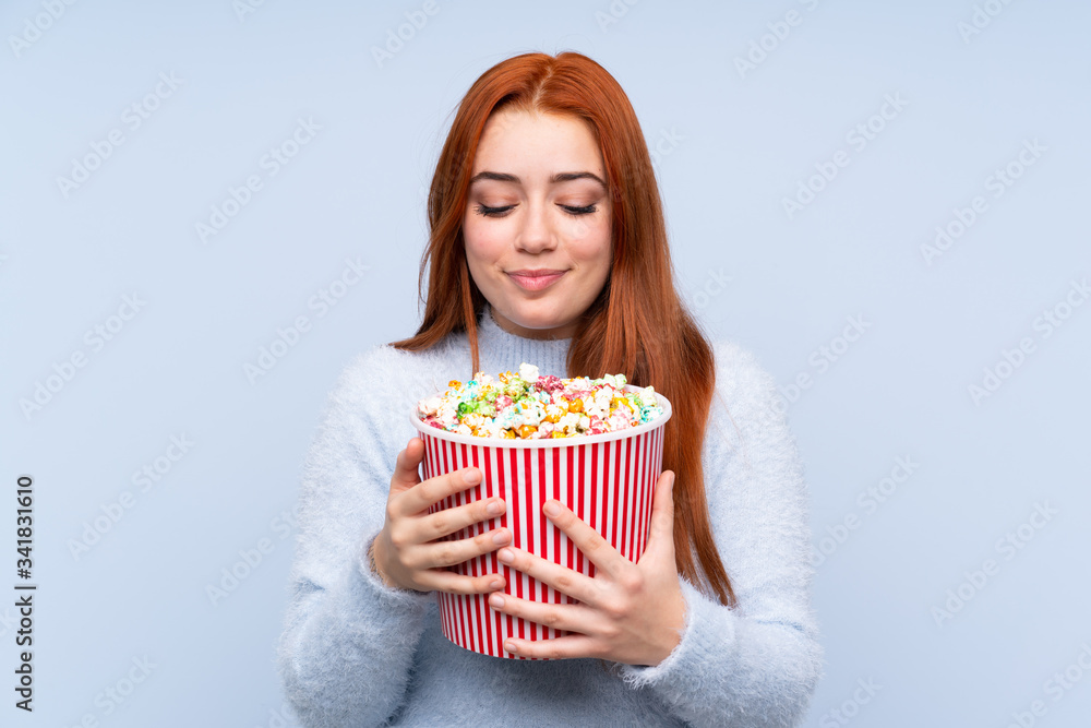 Redhead teenager girl over isolated blue background holding a big bucket of popcorns Redhead teenager girl over isolated blue background holding a big bucket of popcorns