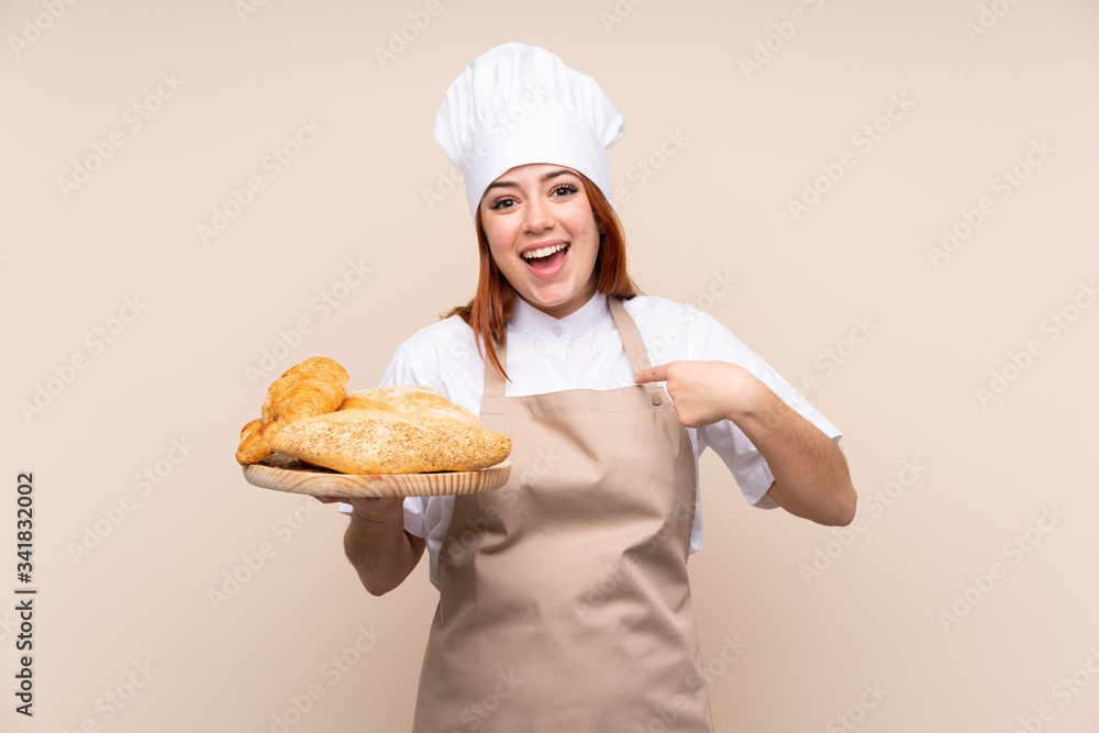 Redhead teenager girl in chef uniform. Female baker holding a table with several breads with surprise facial expression Redhead teenager girl in chef uniform. Female baker holding a table with several breads with surprise facial expression