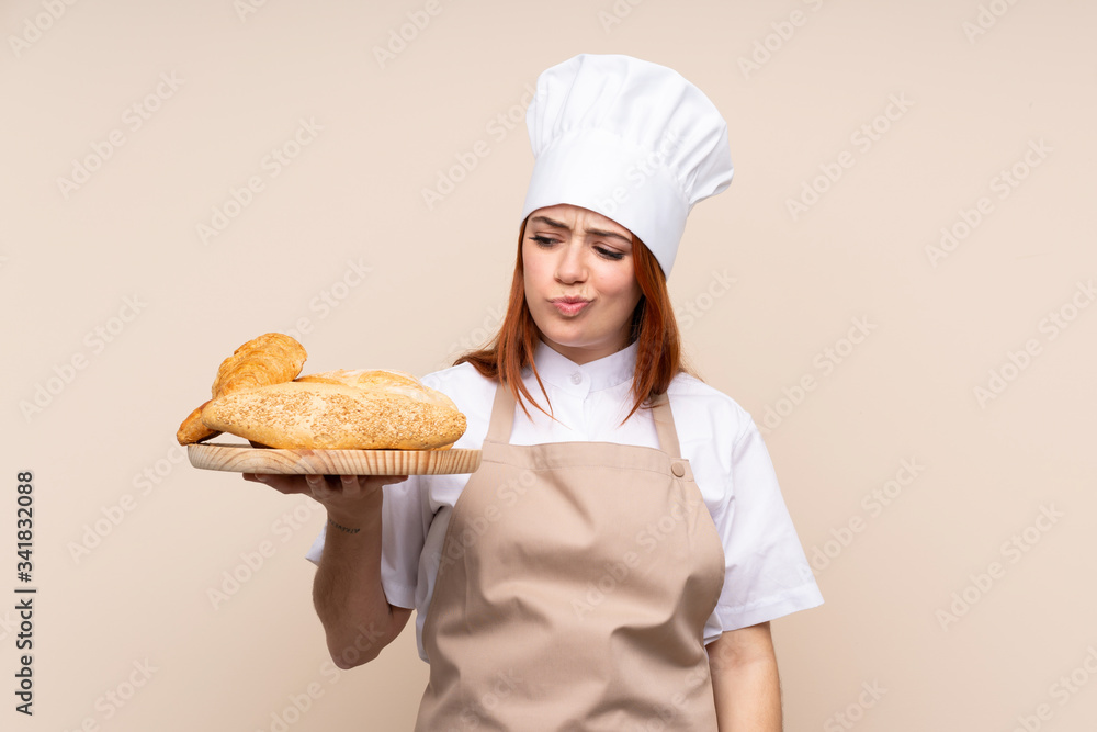 Redhead teenager girl in chef uniform. Female baker holding a table with several breads with sad expression Redhead teenager girl in chef uniform. Female baker holding a table with several breads with sad expression