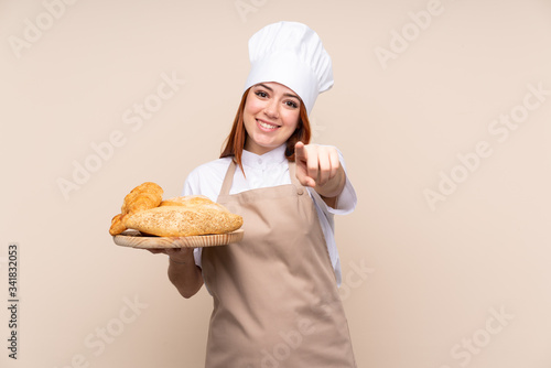 Redhead teenager girl in chef uniform. Female baker holding a table with several breads points finger at you with a confident expression