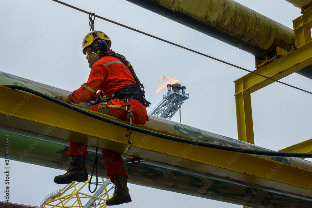 Working at height. An abseiler wearing Personal Protective Equipment (PPE) sitting on the structure for touch up painting activities with background flare tip burning in the sky.