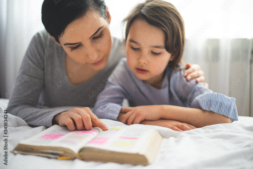 Mother and her daughter reading from bible and praying in their knees near the bed Mother and her daughter reading from bible and praying in their knees near the bed
