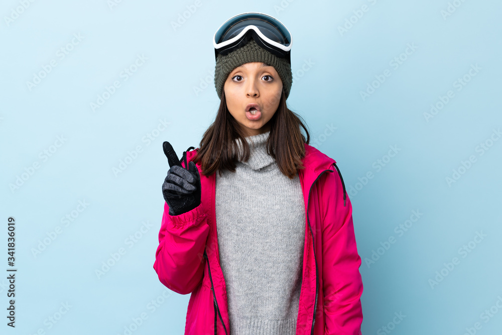 Mixed race skier girl with snowboarding glasses over isolated blue background thinking an idea pointing the finger up. Mixed race skier girl with snowboarding glasses over isolated blue background thinking an idea pointing the finger up.