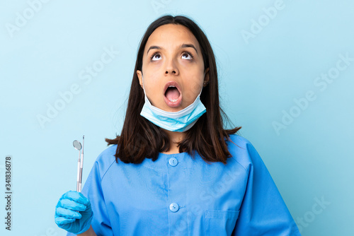 Young brunette mixed race dentist woman holding tools over isolated background looking up and with surprised expression