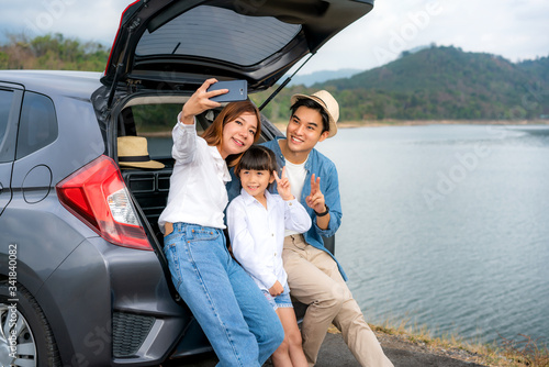 Portrait of Asian family sitting in car with father, mother and daughter selfie with lake and mountain view by smartphone while vacation together in holiday. Happy family time. © ake1150