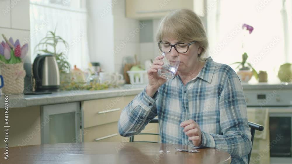 Woman Swallows Pill And Drinks Water While Sitting In Her Kitchen. Woman Swallows Pill And Drinks Water While Sitting In Her Kitchen.