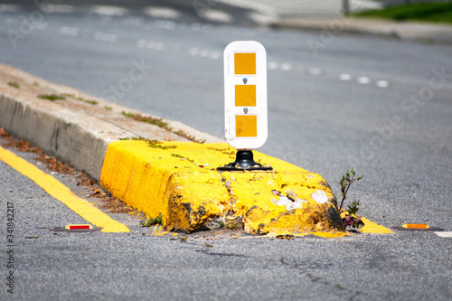 Dividing median curb on road with white and yellow warning reflecting sign on pole. Yellow traffic marking paint an overgrown vegetation