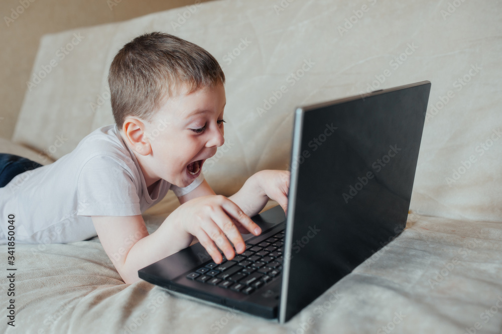 beautiful blond schoolboy boy is lying on the sofa with a laptop, doing homework and shouting at the screen. Remote education during the quarantine period. Online lessons for school children beautiful blond schoolboy boy is lying on the sofa with a laptop, doing homework and shouting at the screen. Remote education during the quarantine period. Online lessons for school children