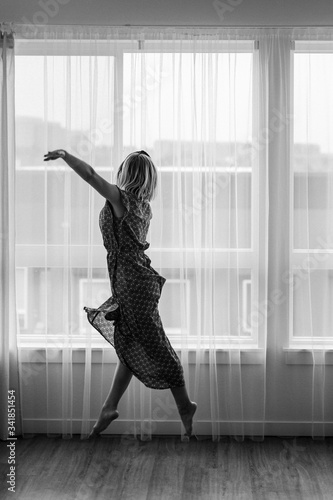 Fototapeta Naklejka Na Ścianę i Meble -  Black and white concept, lady dances in maxi dress in front of the window. Ballerina is dancing. Dance moves. Self isolation stay home coronavirus.Dancer shows neighbors her dancing moves.Large window