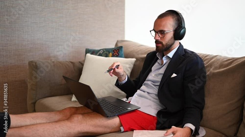 Young man working from home sitting on the sofa with earphones, suit and shorts