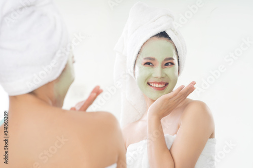 Asian woman is applying facial mask. She is looking in the mirror.