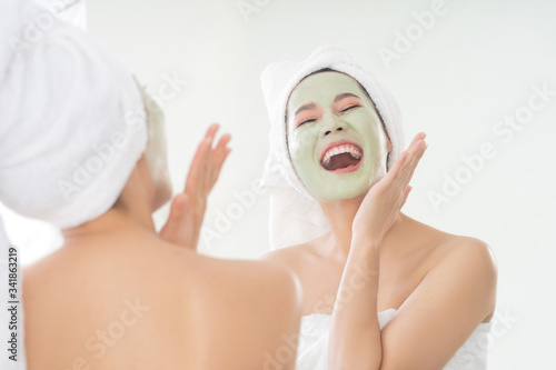 Asian woman is applying facial mask. She is looking in the mirror.