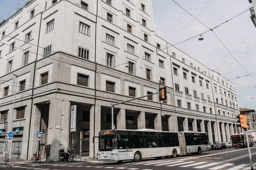 A view of a tall architectural white building with a white bus passing along an empty street and yellow pedestrian lights installed, the city is like new York but this is Italy | BOLOGNA, ITALY - 17 S