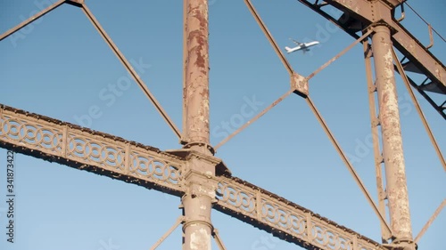 A plane passes from left to right behind a close up rusting Victorian gas holder at gasworks in urban East London made of iron girders. With light blue sky and no cloud photo