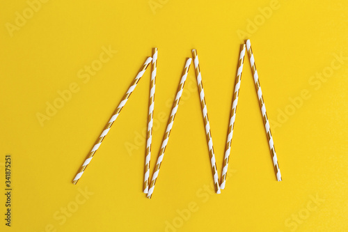 Bright paper drinking straws with white and golden striped on yellow background with copy space. Concept for holiday or celebration. Flat lay.