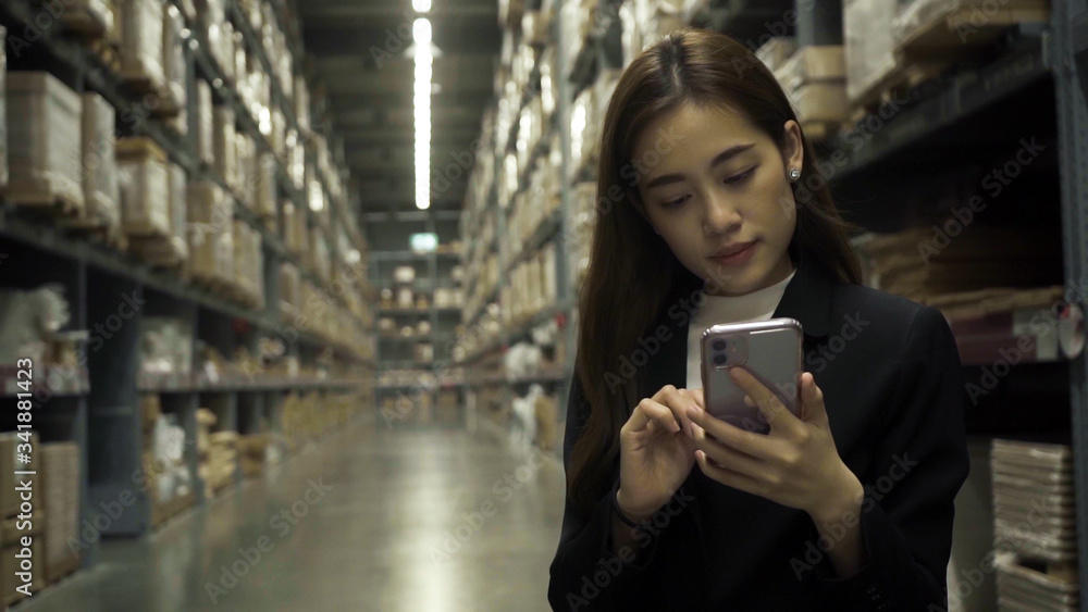Asian female worker wearing a black suit Checking in warehouse For her company To export to customers By using a smartphone to take notes.