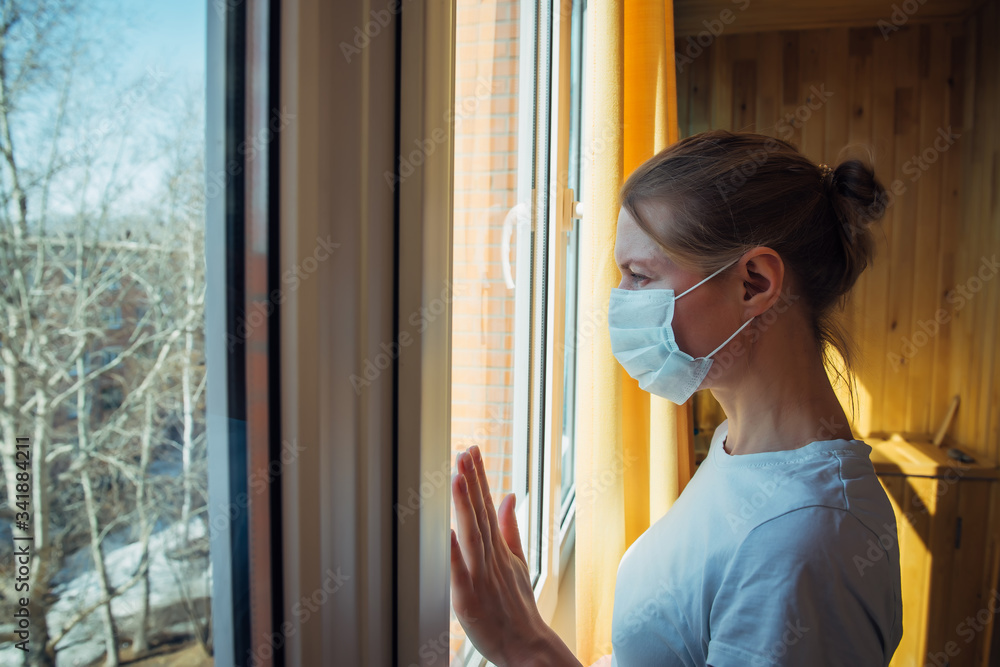 Sad lonely woman in protective medical mask on her face looking at window. Chinese pandemic coronavirus, virus Covid-19. Quarantine, prevent infection, home Isolation, protective against SARS-CoV-2.