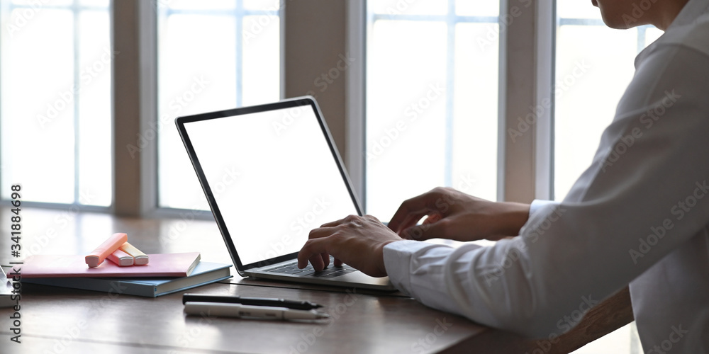 Cropped image of young businessman typing on computer laptop with white blank screen while sitting at the wooden working desk over comfortable living room windows as background. Cropped image of young businessman typing on computer laptop with white blank screen while sitting at the wooden working desk over comfortable living room windows as background.