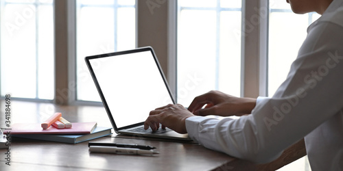 Cropped image of young businessman typing on computer laptop with white blank screen while sitting at the wooden working desk over comfortable living room windows as background.