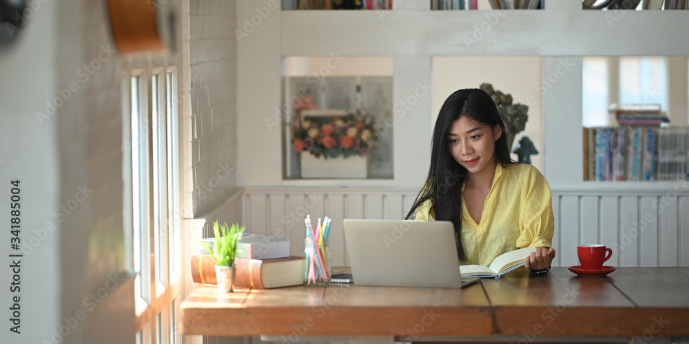 Photo of woman sitting and taking note while sitting in front her computer laptop that putting on wooden working desk and surrounded by coffee cup, pencil holder, potted plant and stack of books Photo of woman sitting and taking note while sitting in front her computer laptop that putting on wooden working desk and surrounded by coffee cup, pencil holder, potted plant and stack of books