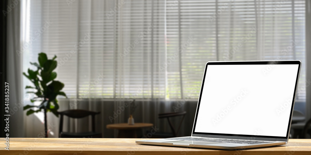 Fototapeta premium Photo of Computer laptop with white blank screen putting on wooden working desk over living room curtain and potted plant as living room background.