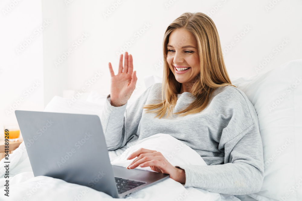 Image of woman waving hand and using laptop while lying in bad Image of woman waving hand and using laptop while lying in bad