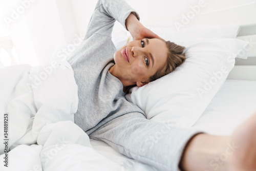 Image of woman smiling and taking selfie while lying in bed