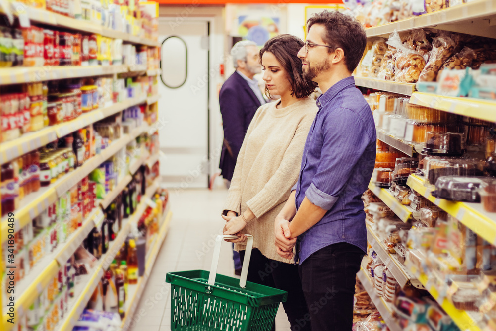 Couple looking at shelves in grocery store. Focused young man and woman holding basket and choosing products in supermarket. Shopping concept