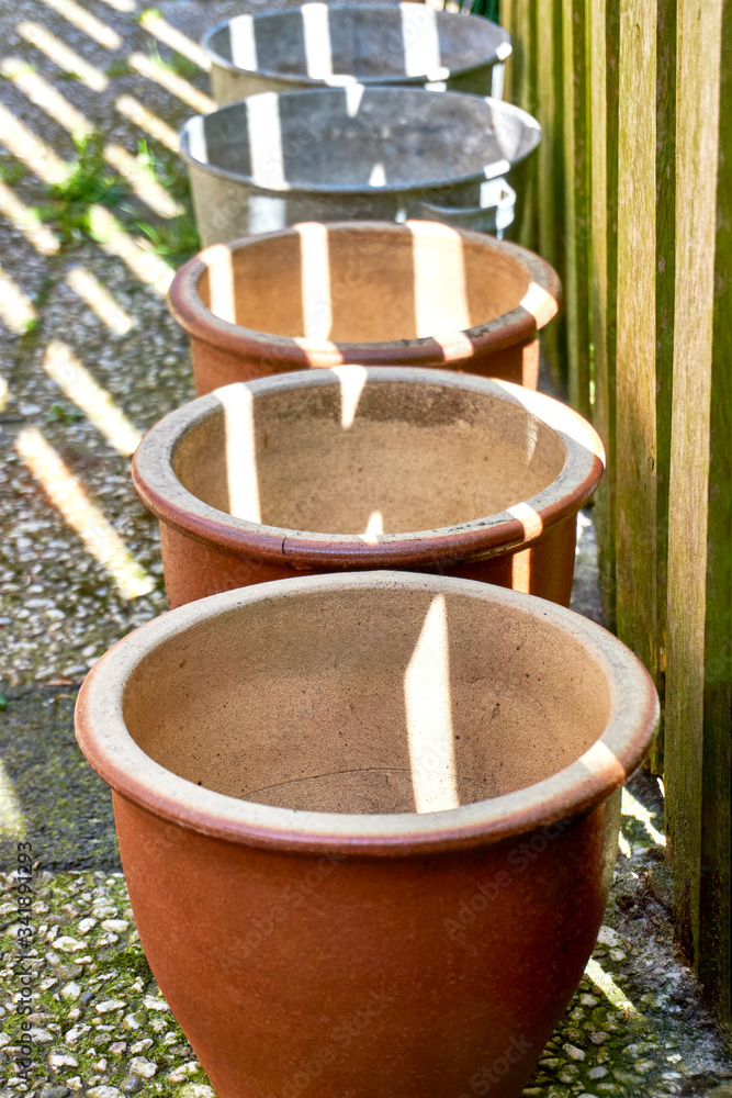 A row of empty zinc and brown ceramic flower pots in a garden with stripes of sunlight shining trough a fence. Vertical image.