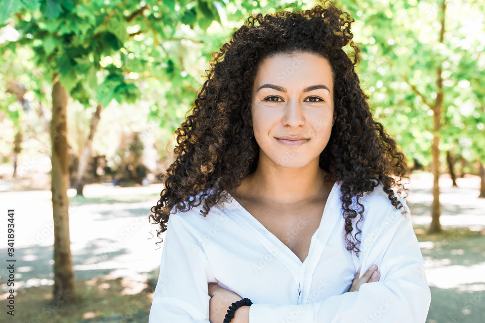 Smiling curly woman with crossed arms looking at camera. Beautiful young lady posing in spring park. Concept of confidence Smiling curly woman with crossed arms looking at camera. Beautiful young lady posing in spring park. Concept of confidence