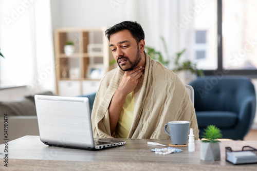 healthcare, technology and people concept - sick indian man in blanket with sore throat having video call on laptop computer at home photo