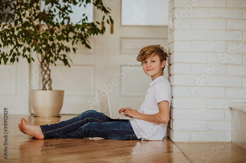 Young  blond boy in white shirt and jeans printing on the white laptop sitting on the floor at home