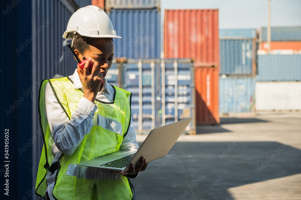 Black foreman woman worker working checking at Container cargo harbor holding laptop computer and walkie-talkie to loading containers. African dock female Logistics import export shipping concept. Black foreman woman worker working checking at Container cargo harbor holding laptop computer and walkie-talkie to loading containers. African dock female Logistics import export shipping concept.