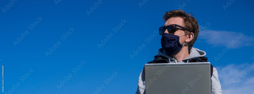 Fototapeta Young man in medical mask works or studies remotely during pandemic on Sunny day sitting on grass in park outdoors. Spring relax of coronavirus epidemic. Urban man with laptop in park.Pollen allergy