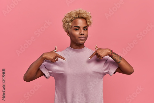 Self assured proud African American woman has blonde hairstyle satisfied with her own high achievements, points with two hands at herself, or at a t-shirt for printing, isolated over pink background photo