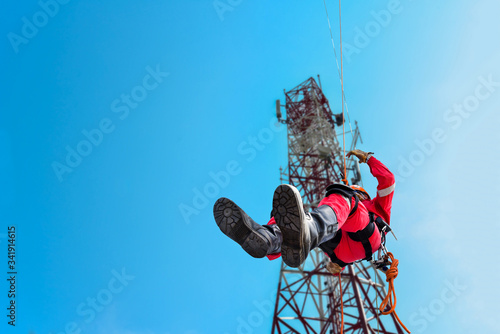 Rope access Worker on abseiling from high places, wearing full safety body harness with helmet protection hanging upside on mast communication background on blue sky. photo