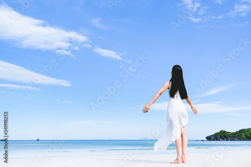 Asian woman, long black hair, wore white dress and hat standing at the beach,arms open and facing back by the sea with copy space of blue sky summer holiday and vacation concept.