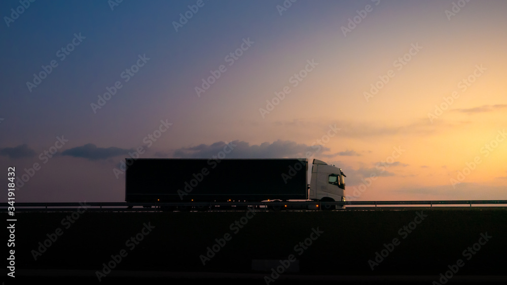 Silhouette of a truck on road at sunset.Semi truck trailer silhouette with setting sun.Delivery truck on asphalt road highway at sunset.Truck passing by a railing. Underexposed pic. Silhouette of a truck on road at sunset.Semi truck trailer silhouette with setting sun.Delivery truck on asphalt road highway at sunset.Truck passing by a railing. Underexposed pic.